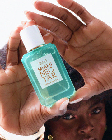 Closeup of woman with brown hair and French manicure wearing white and gold bracelets holding MIAMI NECTAR Eau de Parfum 50ml in both of her hands above her head