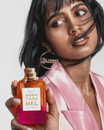 Woman with black hair wearing a pink blazer and gold queen hoop earrings holds QUEENS CARAMEL Eau de Parfum 50ml without cap covered in caramel drizzle on white background. 