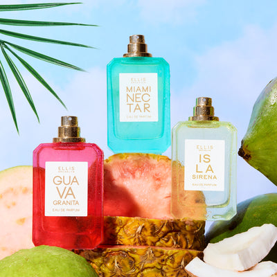 GUAVA GRANITA 50ml Eau de Parfum, MIAMI NECTAR 50ml Eau de Parfum and  ISLA SIRENA 50ml Eau de Parfum without caps covered in water droplets sitting on sliced guavas, sliced pineapple, sliced coconuts, and green coconuts on blue background with green palm leaves | english_image