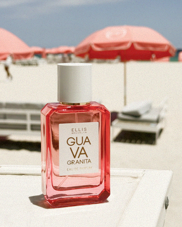 GUAVA GRANITA Eau de Parfum 50ml sits on white table against a beach background of white sand, white lounge chairs, pink umbrellas, and blue sky