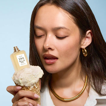 A woman with black hair wearing a white shirt and gold hoop earrings and necklace holds a vanilla ice cream cone with VANILLA SALT Eau de Parfum 7.5ml without cap on top all on a blue background.
