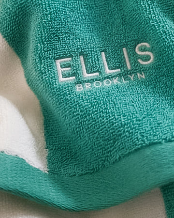 Close up of teal and white striped beach towel embroidered with white copy that reads "Ellis Brooklyn"