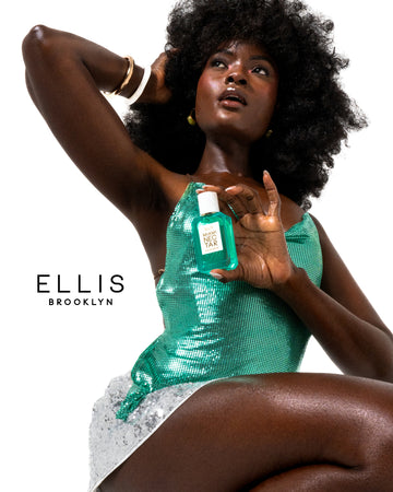 Woman with brown hair wearing a silver sequin miniskirt, teal sequin tank top, and white and gold bracelets sits with her one hand on her head and the other hand in front of her chest holding MIAMI NECTAR Eau de Parfum 50ml against a white background with black text that reads 'Ellis Brooklyn'