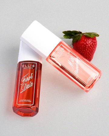 MISS STRAWBERRY Perfume Mist 90ml lays down on FIRST DATE Perfume Mist 90 ml cap next to a strawberry all on a gray background
