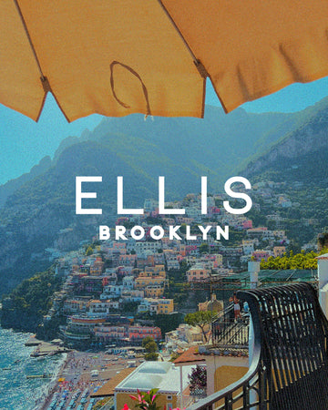 Mountain with colorful buildings against a blue ocean and beach covered in people and umbrellas with white text that reads "Ellis Brooklyn" in the background of a metal balcony and chair and a yellow umbrella 