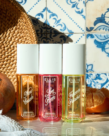 NEW CRUSH Perfume Mist 90ml, LOVE POM Perfume Mist 90ml, and FOREVER PISTACHIO Perfume Mist 90ml between tan basket, pomegranate, partially peeled clementine, and white lace fabric on top of a shiny white surface and in front of white tiles with blue floral designs