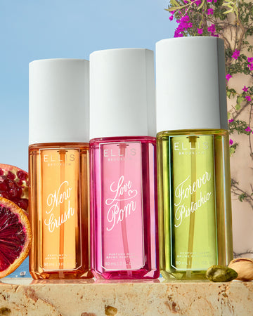 NEW CRUSH Perfume Mist 90ml, LOVE POM Perfume Mist 90ml, and FOREVER PISTACHIO Perfume Mist 90ml stand on tan marble next to a sliced pomegranate, a sliced blood orange, and pistachios in front of a tan wall with purple flowers, a blue ocean, and blue sky 