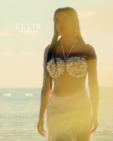 Woman with wet, brown hair wearing a shell bikini top and white skirt stands in front of the ocean and blue sky with golden sunlight streaming in front of her with white copy that reads "Ellis Brooklyn"