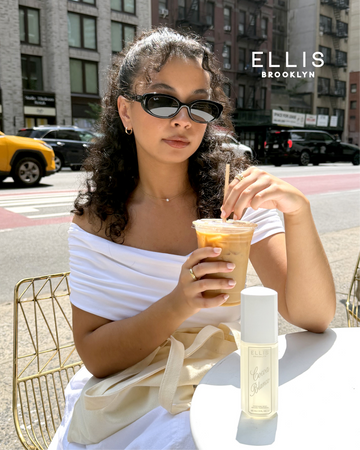 A woman with brown hair wearing black sunglasses and a white off-the-shoulder top sits at an outdoor cafe table holding an iced coffee, with COCOA BLANCO Perfume Mist 90ml on the table in front of her. The background shows a city street with buildings, cars, a yellow cab, and the white Ellis Brooklyn logo.