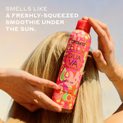 Woman with blonde hair and gold ring and bracelets holds Amika x Ellis Brooklyn GUAVA GRANITA Perk Up Dry Shampoo against the back of her head against blue and yellow sky with white text that reads " smells like a freshly-squeezed smoothie under the sun"