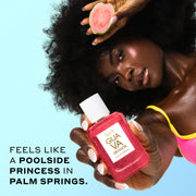 Woman with brown hair wearing a pink and yellow bikini top holds GUAVA GRANITA Eau de Parfum 50ml covered in water droplets out in front of her in one hand and a slice of guava in her other hand next to her head against a blue background with black text that reads "Feels like a poolside princess in Palm Springs."