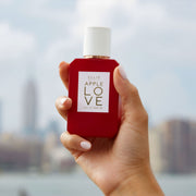 Modèle avec du vernis à ongles rose clair tenant un flacon Apple Love 50 ml Eau de Parfum avec la ville en arrière-plan.