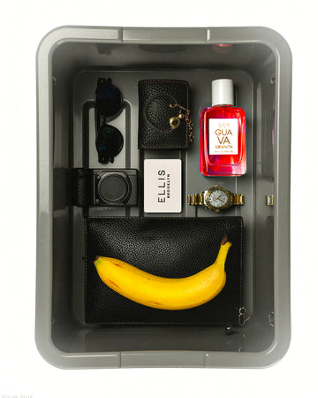 Gray bin on a white background holds a black pair of folded sunglasses, a black purse with a gold chain, Guava Granita Eau de Parfum 50ml, a black digital camera, a white Ellis Brooklyn card, a gold watch, and a black clutch with an unpeeled banana on top of it