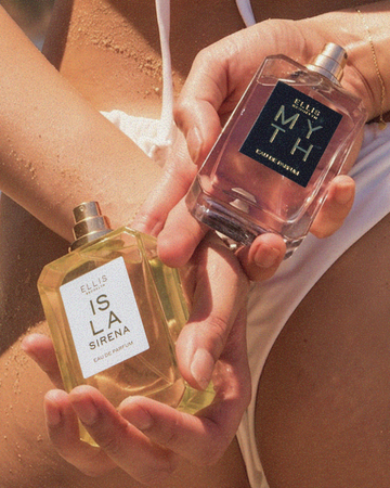 Closeup of woman wearing white bikini and gold bracelets with sand on her holding ISLA SIRENA Eau de Parfum 50ml without cap and MYTH Eau de Parfum 50ml without cap behind her back 