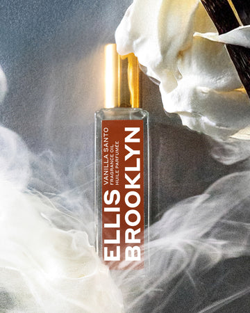Vanilla Santo Fragrance Oil 15ml near whipped cream swirl and vanilla beans surrounded by smoke, on grey background.