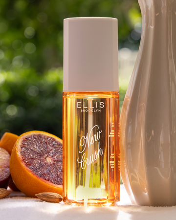 NEW CRUSH Perfume Mist on white table next to halved blood oranges, a white vase, and almonds with green leaves in the background