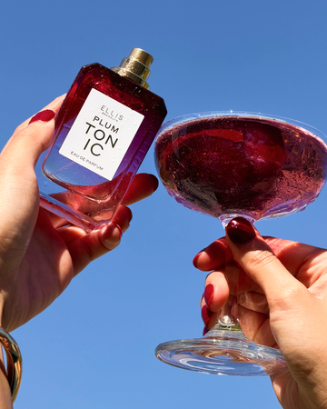 Hands with red manicure hold PLUM TONIC Eau de Parfum 50ml without cap and a clear coupe glass filled with purple liquid against a blue sky.