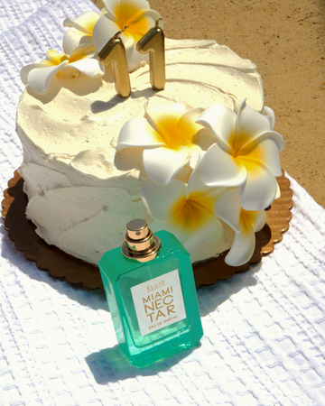 MIAMI NECTAR Eau de Parfum 50ml without cap stands next to white cake with white and yellow flowers and gold eleven candles on a gold plate sitting on white textured blanket next to tan sand