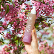 IMAGE 5: MODEL HOLDING  SAND FRAGRANCE AND BODY MIST 100ML NEAR FLOWERS