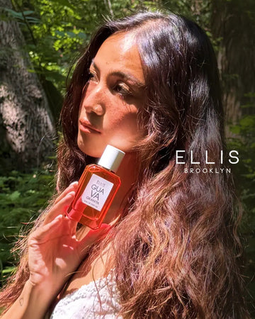Model with brown hair wearing white top holding Guava Granita 50ml Eau De Parfum to face in woods, with Ellis Brooklyn logo.