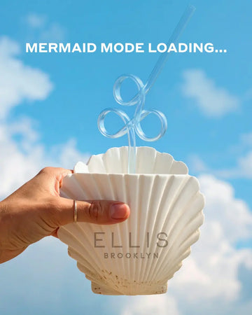 hand with gold ring on thumb holding shell shaped cup with Ellis Brooklyn logo and clear bendy straw against sky with clouds with white text that reads 