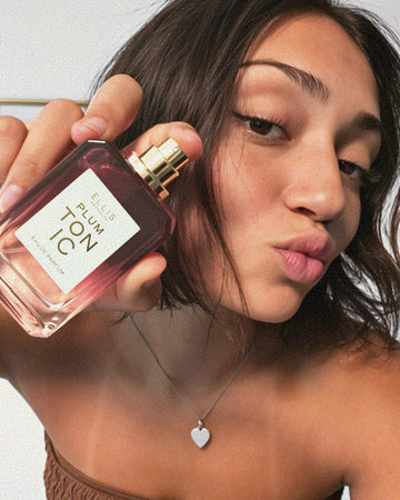 Woman with brown hair wearing a silver heart necklace and brown strapless top holds PLUM TONIC Eau de Parfum 50ml without cap against her cheek on a white background