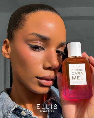 Model with brown hair wearing denim jacket holding Queens Caramel 50ml Eau De Parfum in one hand near face, with white room in background.