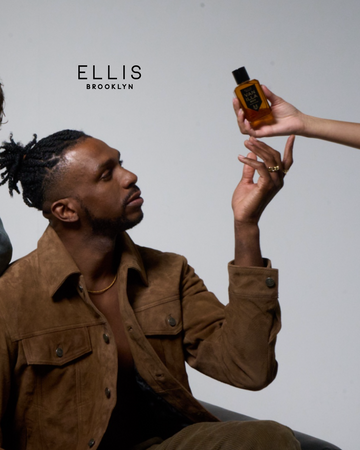 Model with brown hair wearing brown suede jacket, brown pants,  gold rings and gold necklace being handed Vanilla Man 50ml Eau De Parfum on grey background with black Ellis Brooklyn logo.