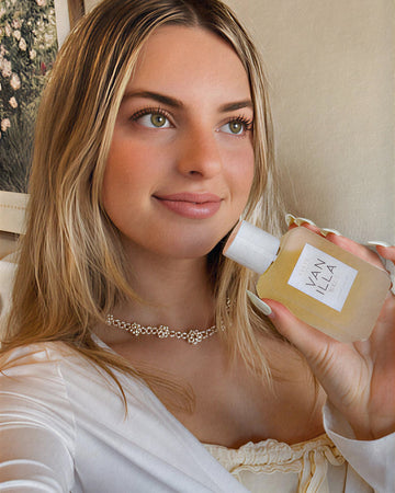 Model with long, blonde hair in yellow shirt, white sweater, and floral necklace holds Vanilla Salt Eau de Parfum 50ml while looking off to the side against a beige wall