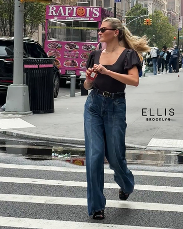 Model with brown hair wearing black shirt, blue jeans, black shoes, and brown sunglasses walking across street in New York City while holding Queens Caramel 50ml Eau De Parfum with both hands.