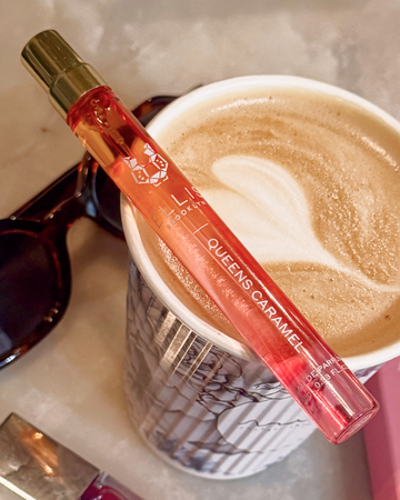 Queens Caramel 10ml Eau De Parfum Travel Spray resting on top of gray floral print coffee cup on marble table next to lip gloss, sunglasses, and pink book.