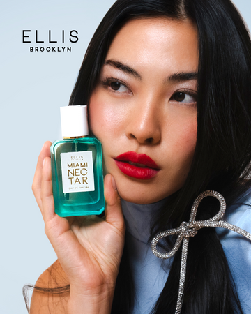 Model with brown hair wearing red lipstick, blue dress, and diamond clip holding Miami Nectar 50ml Eau De Parfum to face, on blue background with black Ellis Brooklyn logo.