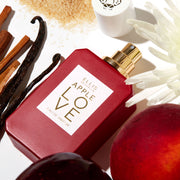 Apple Love 50ml Eau de Parfum avec le bouchon retiré à côté d'une fleur blanche, de pomme, de prune, de bois de santal, de gousses de vanille et de sucre sur fond blanc.