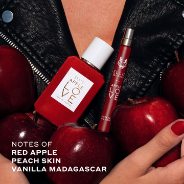 Model with red nails wearing black leather jacket holding red apples, Apple Love 50ml Eau De Parfum and Apple Love 10ml Eau De Parfum Travel Spray in hands agaisnt chest. 