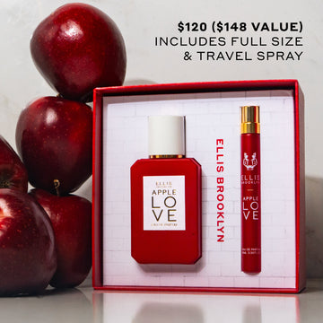 Apple Love Eau De Parfum Gift Set Box showing Apple Love 50ml Eau De Parfum and Apple Love 10ml Eau De Parfum Travel Spray on white table next to stack of red apples on white background with black text that reads "$120 ($148 value) includes full size & travel spray"