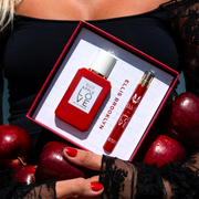 Model with red nails and blonde hair wearing black shirt with black lace sleeves holding Apple Love Eau De Parfum Gift set box showing Apple Love 50ml Eau De Parfum and Apple Love 10ml Eau De Parfum and red apples in arms to stomach.