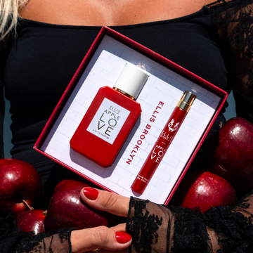 Model with red nails and blonde hair wearing black shirt with black lace sleeves holding Apple Love Eau De Parfum Gift set box showing Apple Love 50ml Eau De Parfum and Apple Love 10ml Eau De Parfum and red apples in arms to stomach.
