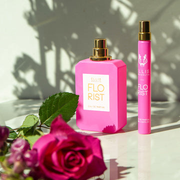 Flacon de parfum Florist 50 ml avec le bouchon enlevé sur une table blanche à côté du spray de voyage Florist 10 ml derrière des roses roses, sur fond blanc avec des ombres.