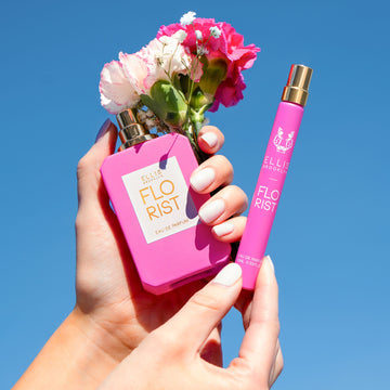 Mannequin avec du vernis à ongles nude tenant le parfum Florist 50ml Eau De Parfum sans le bouchon avec des fleurs roses et blanches dans une main et le spray de voyage Florist 10ml Eau De Parfum dans l'autre main, sur fond bleu.