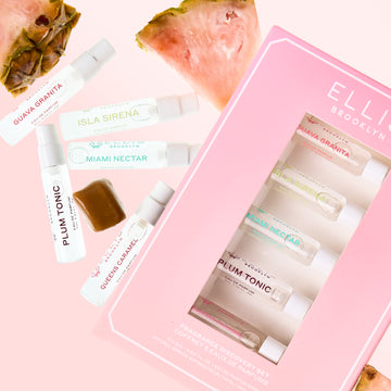Best Sellers Discovery Set next to sliced pink pineapple, a caramel cube, and 2ml vials of GUAVA GRANITA Eau de Parfum, ISLA SIRENA Eau de Parfum, MIAMI NECTAR Eau de Parfum, PLUM TONIC Eau de Parfum and QUEENS CARAMEL Eau de Parfum on a pink background