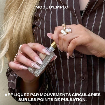 Mannequin aux cheveux blonds et aux ongles vernis de couleur nude, portant une chemise à manches longues rayée marron et blanc ainsi que des bagues en perles assorties ; elle retire le bouchon d'un flacon d'huile parfumée « Blond Musk » (15 ml) sur fond beige, entre des inscriptions en blanc indiquant : « Mode d'emploi : Appliquer sur les points de pulsation ».