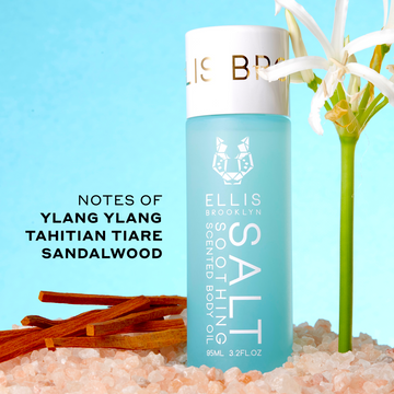 SALT 95ml Soothing Scented Body Oil in pink salt next to white flower and sandalwood with black text that reads "Notes of Ylang Ylang, Tahitian Tiare, Sandalwood" on blue background.
