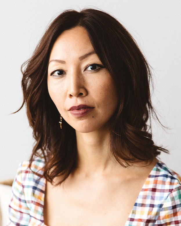 A close-up portrait of Ellis Brooklyn founder Bee Shapiro with shoulder-length brown hair, wearing a multicolor plaid top and a small gold dangle earring, set against a plain light gray background.