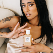 Un mannequin aux cheveux noirs, vernis à ongles blanc, piercings au visage et tatouage de dragon sur le bras porte un débardeur en satin beige en étant allongé sur un canapé blanc cassé et tient le parfum Vanilla Milk 50ml Eau De Parfum à deux mains.