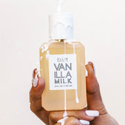 Modèle avec du vernis à ongles blanc tenant le Vanilla Milk 50ml Eau De Parfum avec du lait versé sur la bouteille et la main sur fond blanc.