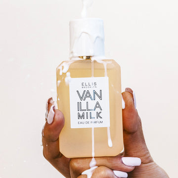 Modèle avec du vernis à ongles blanc tenant le Vanilla Milk 50ml Eau De Parfum avec du lait versé sur la bouteille et la main sur fond blanc.