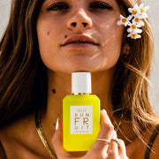 Mannequin aux cheveux bruns et au vernis à ongles blanc portant un débardeur marron, des boucles d'oreilles en or, des colliers en or, des bagues en or et des fleurs blanches dans les cheveux tient le parfum Sun Fruit 50ml sous le menton