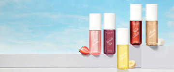 MISS STRAWBERRY Perfume Mist 90ml next to a halved strawberry, VANILLA SIN Perfume Mist 90ml, FIRST DATE Perfume Mist 90ml, and MARSHMALLOWS Perfume Mist 90ml next to a marshmallows all sit on a white platform above BANANA MILKSHAKE Perfume Mist 90ml next to banana slice in front of a blue sky with white clouds. 