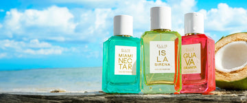 MIAMI NECTAR Eau de Parfum 50ml, ISLA SIRENA Eau de Parfum 50ml, and GUAVA GRANITA Eau de Parfum 50ml all covered in water droplets stand next to half of a coconut on top of gray wood against a background of blue ocean and blue sky with white clouds 