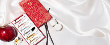 Red An Ellis Brooklyn Mystery Fragrance Discovery Set box opened to show inside and Apple Love 1.5ml Eau De Parfum Vial, Florist 1.5ml Eau De Parfum Vial, Salt 1.5ml Eau De Parfum Vial, Myth 1.5ml Eau De Parfum Vial, Vanilla Milk 1.5ml Eau De Parfum Vial and Sweet 1.5ml Eau De Parfum Vial on white satin sheet with 2 vanilla beans, 2 marshmallows, red apple and white flower.